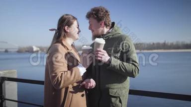 在河岸边爱喝咖啡的年轻夫妇的肖像。 快乐的白人男女在阳光明媚的户外约会
